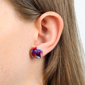A close-up of a person's ear wearing a round stud earring with a multicolor marbled pattern of purple, red, blue, and white.