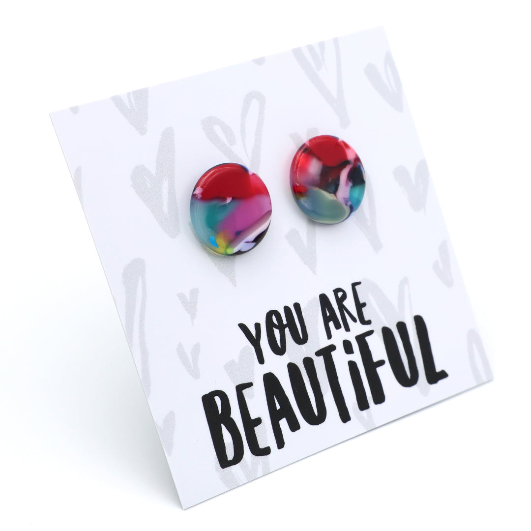 A pair of round stud earrings with a colorful marbled pattern in red, blue, pink, and green, displayed on a white backing card that reads 'YOU ARE BEAUTIFUL'.