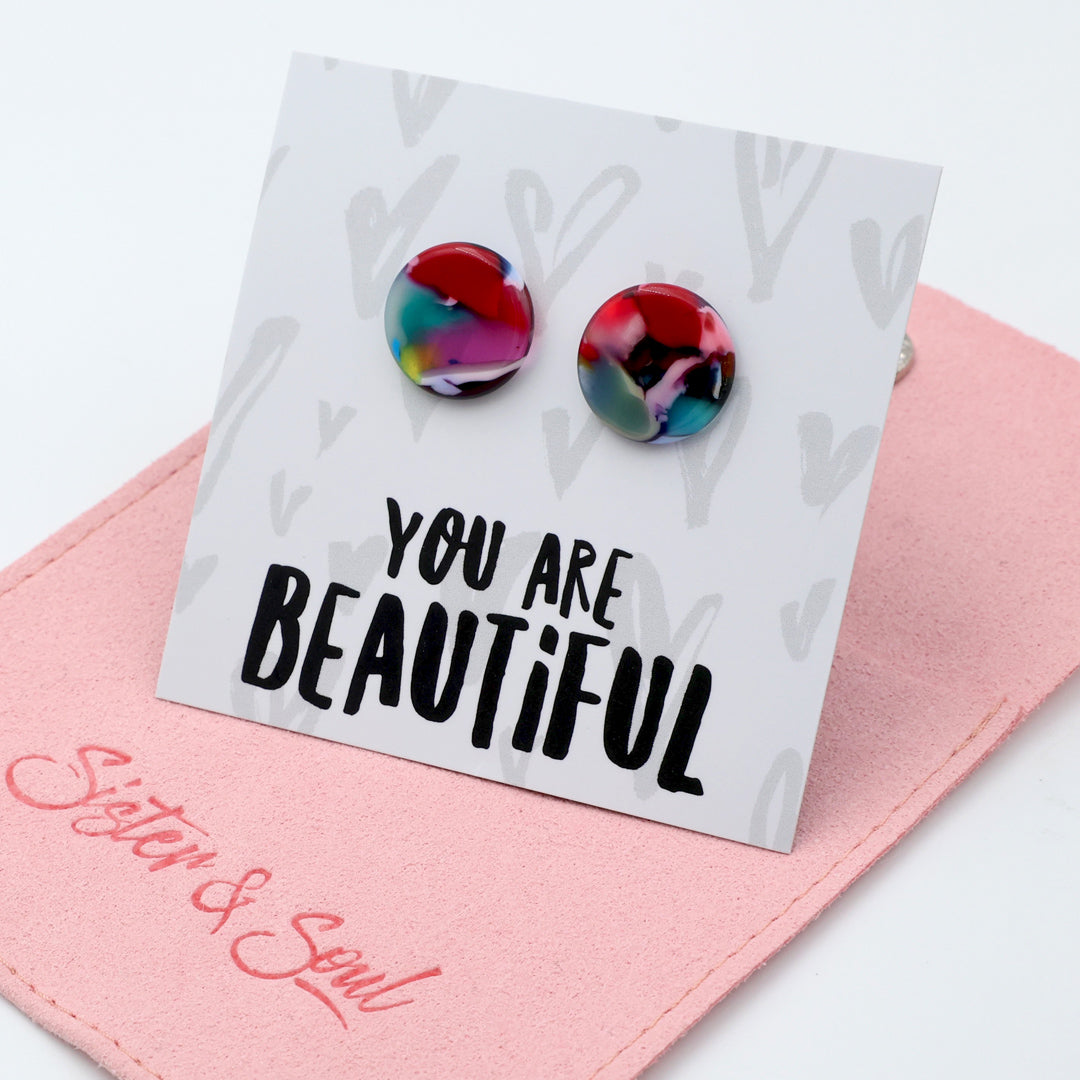 A pair of round stud earrings with a marbled multicolor design in red, blue, green, and purple, displayed on a white backing card that reads 'YOU ARE BEAUTIFUL'. The card is resting on a pink fabric pouch with 'Sister & Soul' written in pink script.