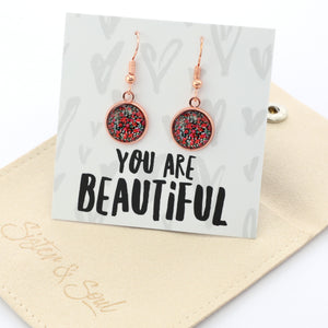 Rose gold earrings on a card with 'You are Beautiful' text, placed on a beige surface.