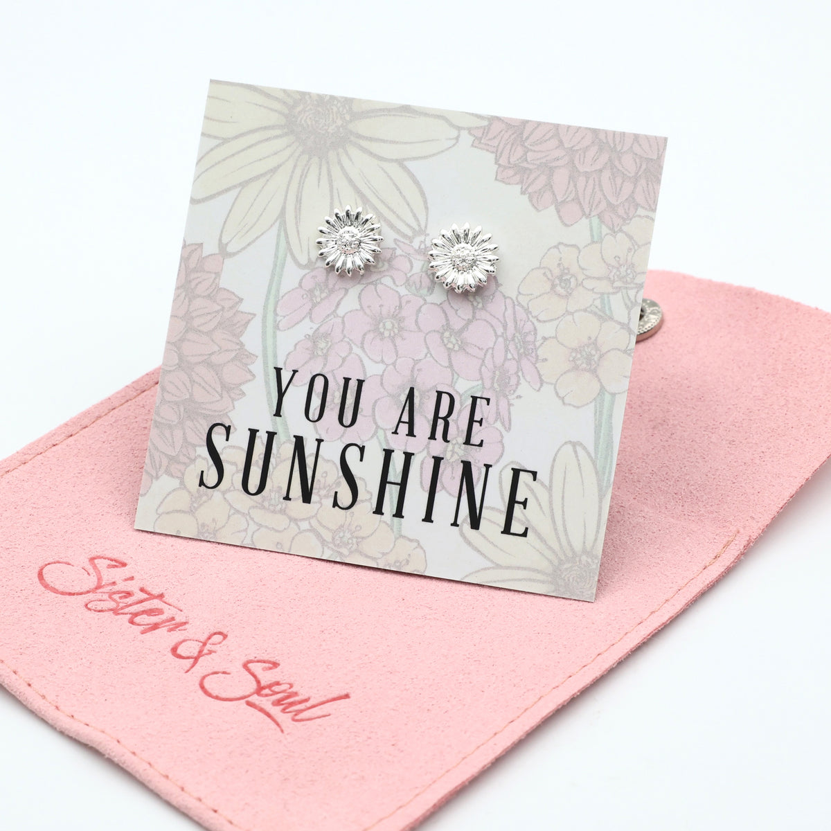 Silver earrings on a card with 'You are Sunshine' text, placed on a pink fabric surface.