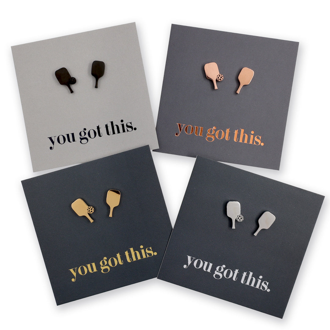 Four square cards arranged on a white background, each with a small pair of pickle ball paddle-shaped studs and the phrase 'you got this.' in different colors: black, rose gold, gold, and silver.