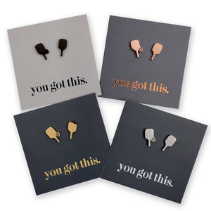 Four square cards arranged on a white background, each with a small pair of pickle ball paddle-shaped studs and the phrase 'you got this.' in different colors: black, rose gold, gold, and silver.