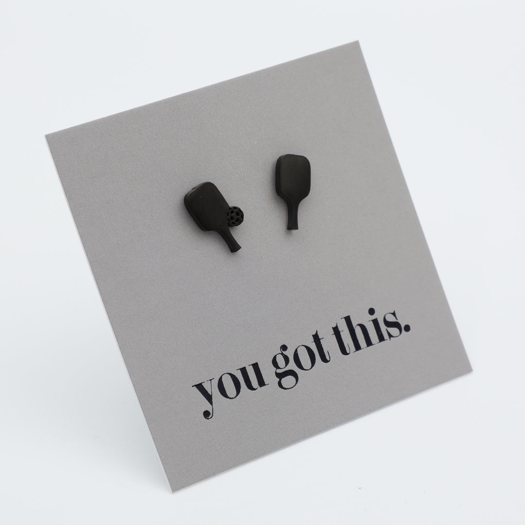 A pair of matte black pickle ball paddle-shaped studs mounted on a light gray square display card with the printed phrase 'you got this.' in black lowercase serif text, photographed on a clean white background.