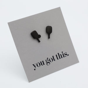 A pair of matte black pickle ball paddle-shaped studs mounted on a light gray square display card with the printed phrase 'you got this.' in black lowercase serif text, photographed on a clean white background.