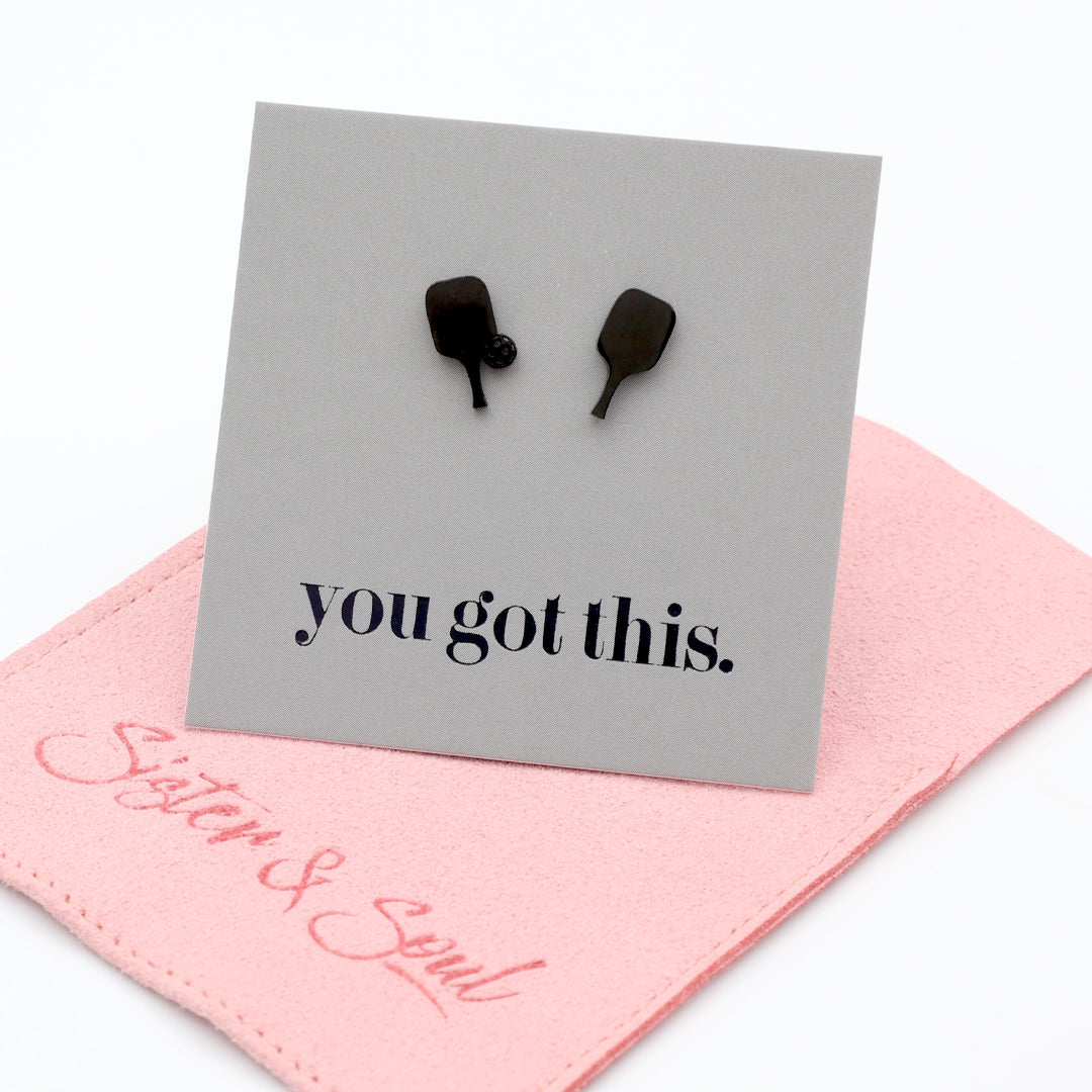 Black pickle ball paddle-shaped stud earrings displayed on a gray card that reads 'you got this.' with a pink fabric pouch underneath on a white background.