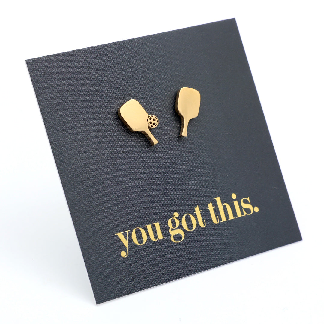 A pair of gold pickle ball paddle-shaped stud earrings displayed on a black square backing card that reads 'you got this.' in gold lowercase text, photographed on a white background.
