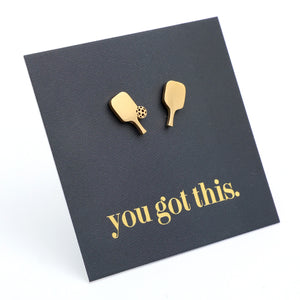 A pair of gold pickle ball paddle-shaped stud earrings displayed on a black square backing card that reads 'you got this.' in gold lowercase text, photographed on a white background.