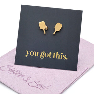 A pair of gold pickle ball paddle-shaped stud earrings displayed on a black backing card that reads 'you got this.' in gold lettering, resting on a purple velvet jewelry pouch embroidered with 'Sister & Soul'.