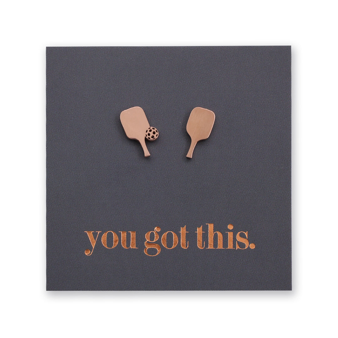 A square dark gray card or plaque with two small metallic rose gold-toned pickleball paddle shapes near the top and the phrase 'you got this.' in matching metallic lettering near the bottom, shown on a white background.