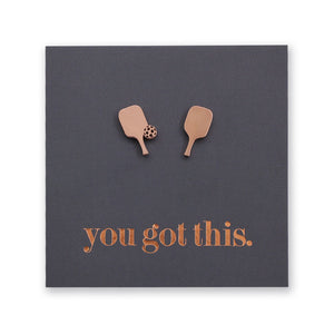 A square dark gray card or plaque with two small metallic rose gold-toned pickleball paddle shapes near the top and the phrase 'you got this.' in matching metallic lettering near the bottom, shown on a white background.