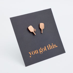 A pair of rose gold stud earrings shaped like pickle ball paddles, displayed on a dark gray textured card that reads 'you got this.' in rose gold lettering, set against a clean white background.