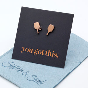 A pair of rose gold stud earrings shaped like pickle ball paddle g, displayed on a black backing card that reads 'you got this.' A light blue fabric pouch with 'Sister & Soul' branding is placed underneath on a white background.
