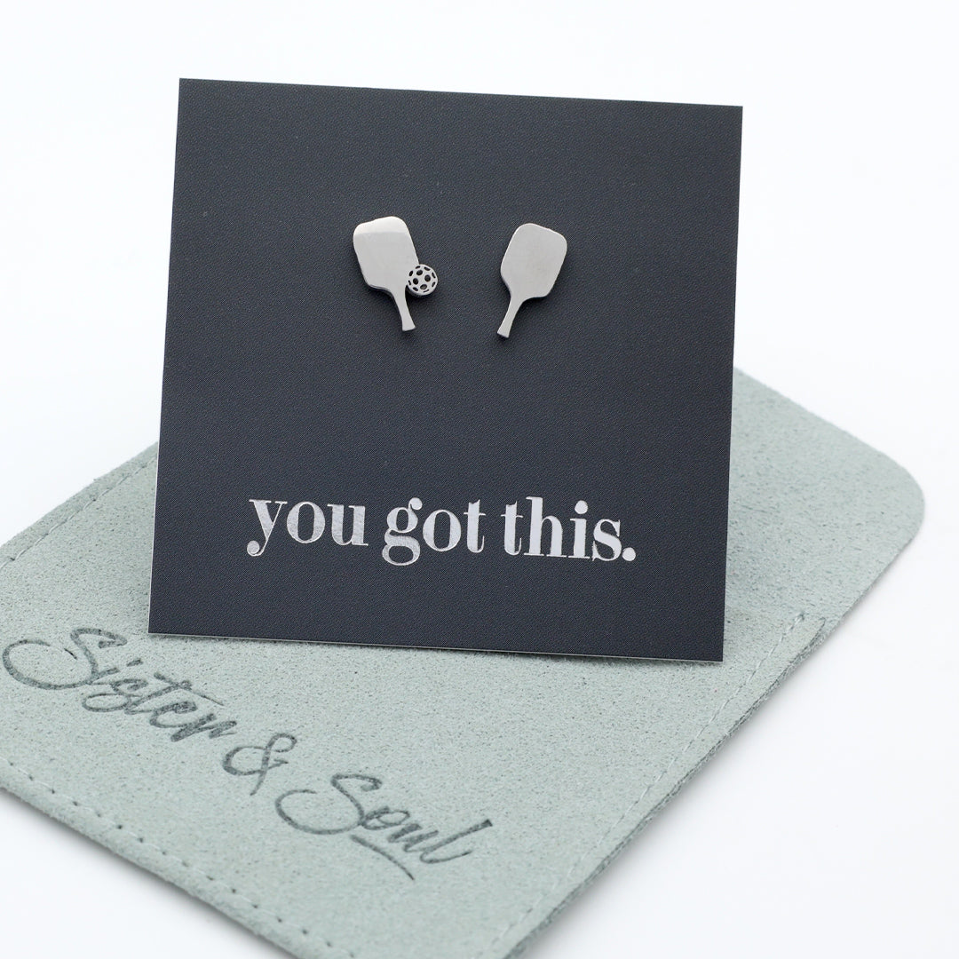 A pair of silver pickle ball paddle-shaped stud earrings displayed on a black backing card that reads 'you got this.' The card is placed on top of a light sage green fabric pouch embroidered with 'Sister & Soul'.