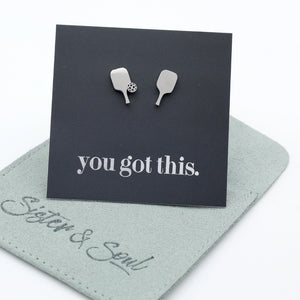 A pair of silver pickle ball paddle-shaped stud earrings displayed on a black backing card that reads 'you got this.' The card is placed on top of a light sage green fabric pouch embroidered with 'Sister & Soul'.