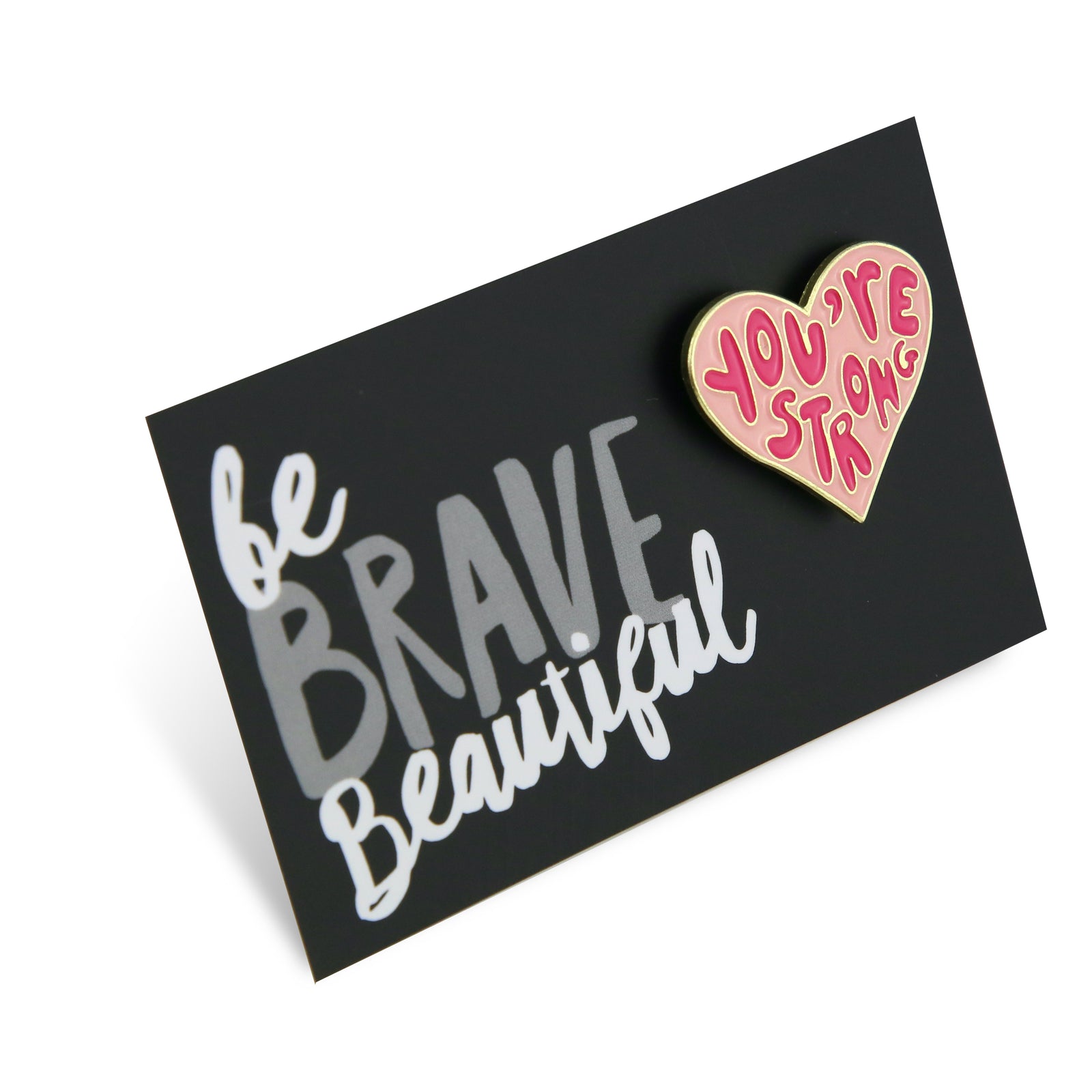 An enamel badge pin with a black background featuring white text 'be BRAVE Beautiful' and a pink heart with the phrase 'YOU'RE STRONG'.