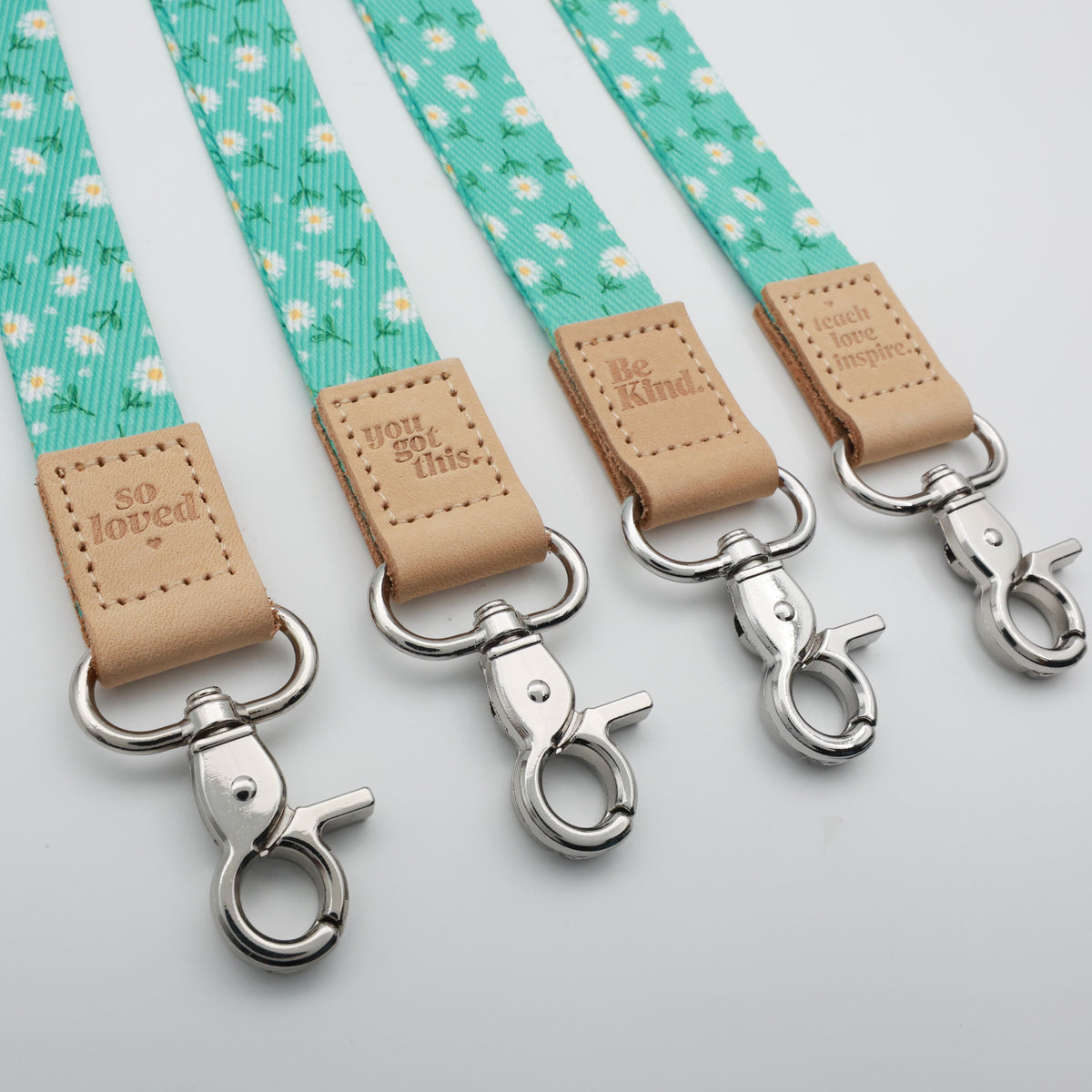 Angled close-up of Aqua Daisies lanyards with tan So Loved, You Got This, Be Kind and Teach Love Inspire tabs and silver-tone clasps.