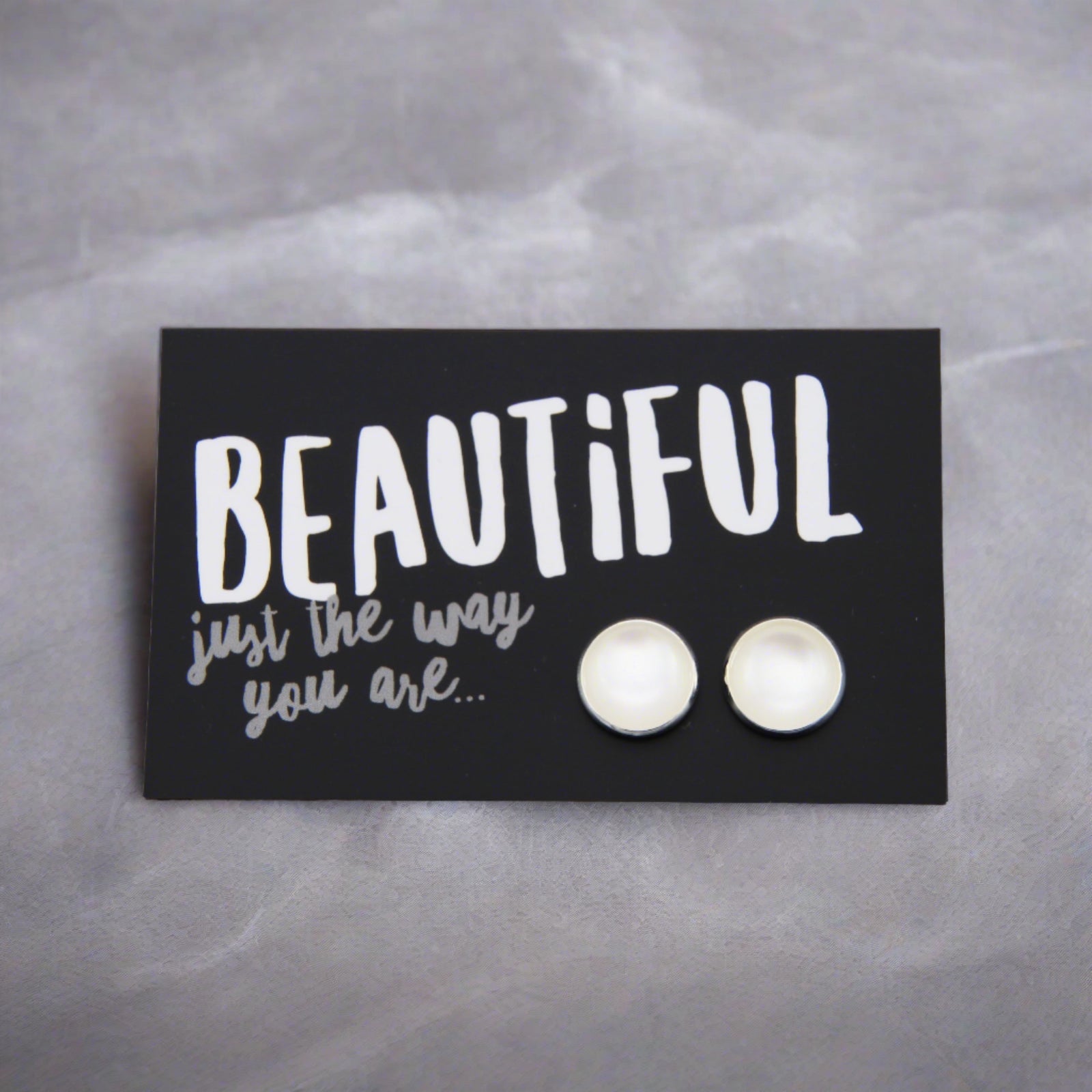 White pearl coloured circle shaped earring studs with silver surround presented on a card that says 'Beautiful just the way you are' 